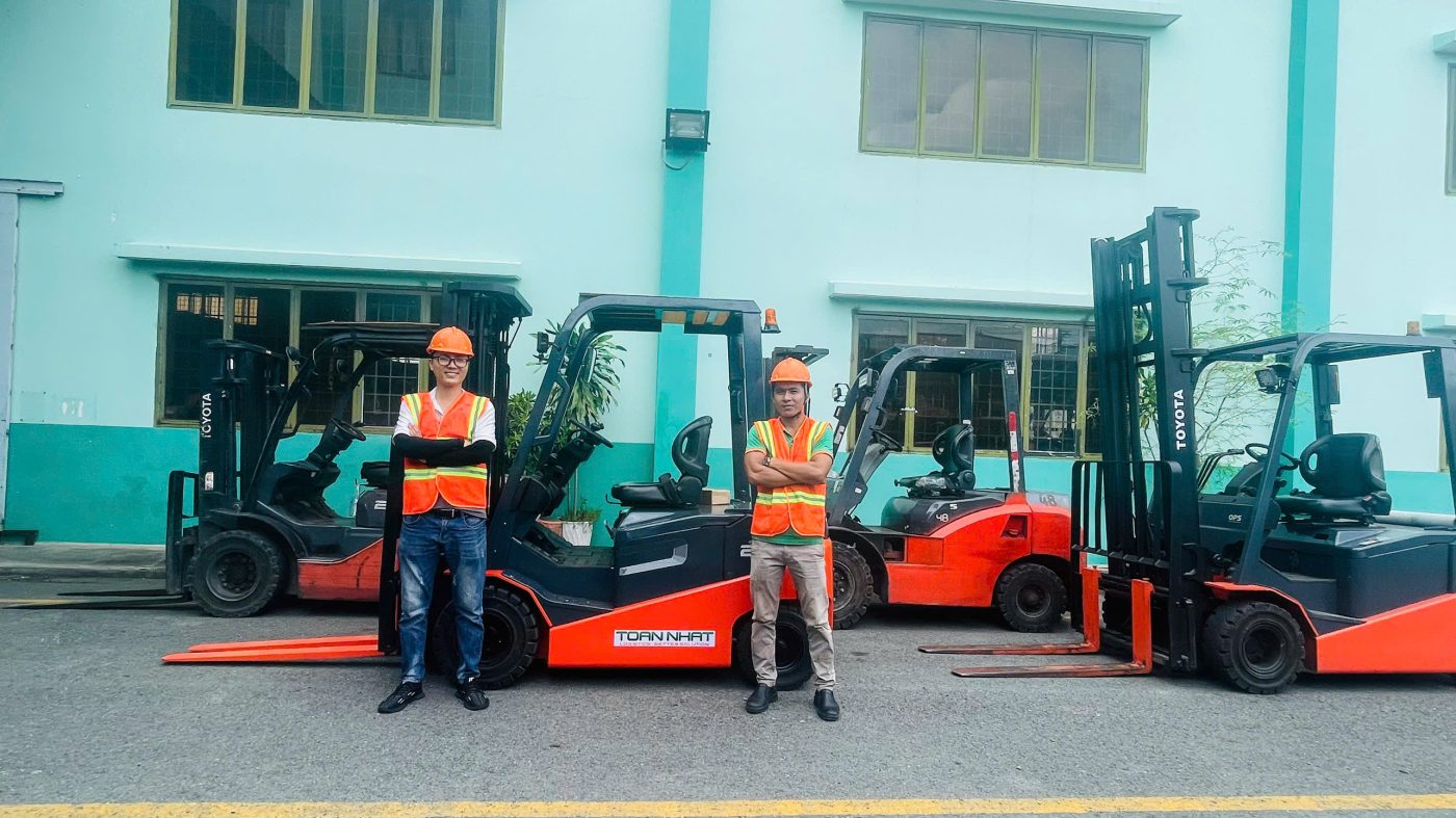 Hai nhân viên của Toàn Nhất Logistics mặc đồ bảo hộ an toàn, đứng tự tin trước dàn ba xe nâng hàng Toyota mới của công ty trong khu vực sân kho.