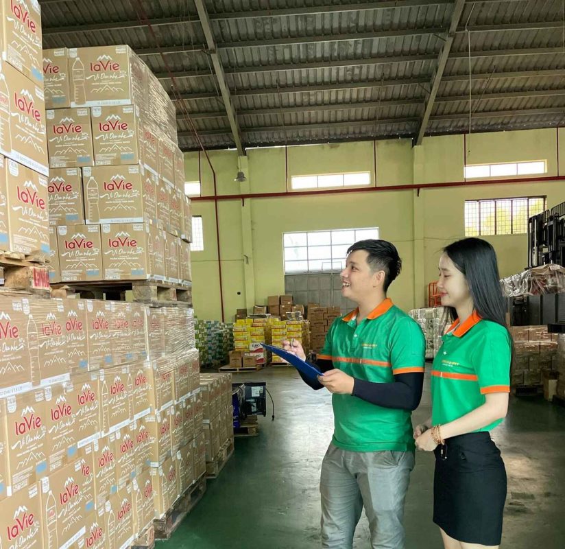 Hai nhân viên Toàn Nhất Logistics mặc đồng phục xanh, đang kiểm tra hàng hóa nước Lavie trong kho, một người cầm bảng kẹp giấy.